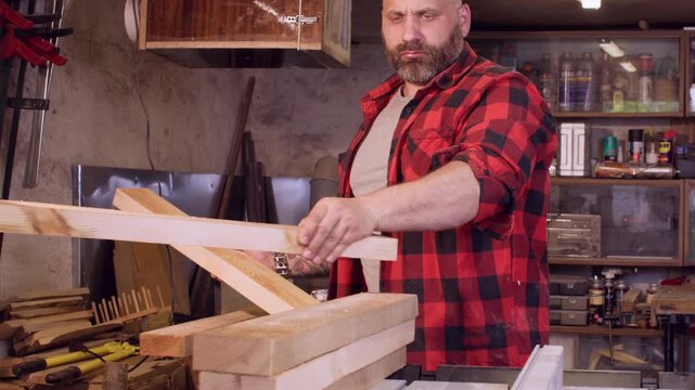 Carpentry service. Stacking wood. Bearded joiner placing freshly cut blocks on table saw in garage workshop with tools set on wall.