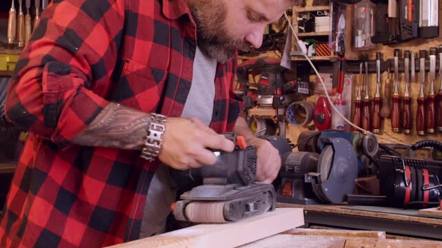 Woodworker service. Joinery process. Man smoothing board surface with electric belt sander in workshop interior with tools kit on shelves.