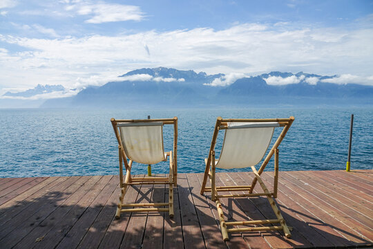 View of two bamboo deck chairs on a wooden pier overlooking Lake Geneva with distant mountains under a blue sky in Vevey, Vaud, Switzerland.