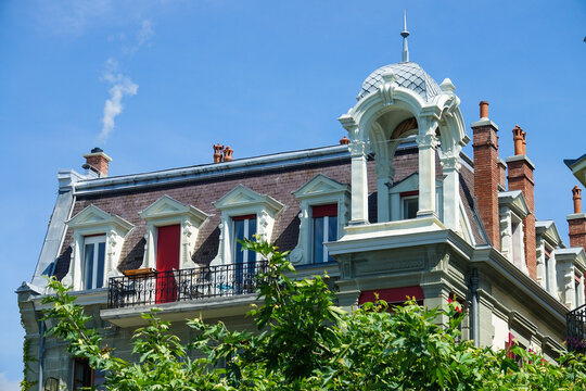 View of a historic building roofline with ornate dormer windows, a small balcony with a red door, and a decorative turret under a clear blue sky in Vevey, Vaud, Switzerland.