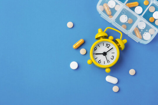 Medicine pills, tablets and capsules with yellow alarm clock and pill box on blue background, top view