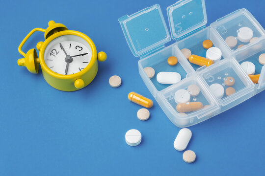 Medicine pills, tablets and capsules with open pill organizer box and yellow alarm clock and  on blue background