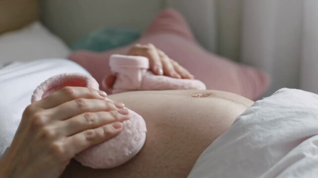 Close-up of a pregnant woman&rsquo;s belly with a pair of tiny pink baby booties placed on top, wearing a white shirt and lying on a bed with soft pillows