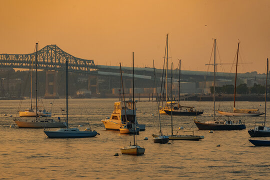 View of sailboats moored in the South Boston Waterfront harbor during a golden sunset with the Tobin Bridge visible in the background, Boston, Massachusetts, United States.