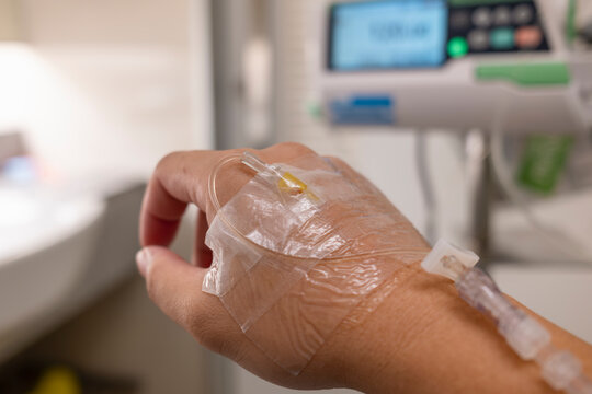 Patient hand with IV drip fluid line close up in hospital room with blurred digital infusion pump monitor background. Healthcare medical insurance policy, illness treatment, and care concept.