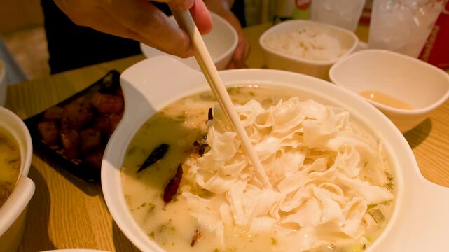Person Adding Wide Rice Noodles to Spicy Fish Soup with Chopsticks