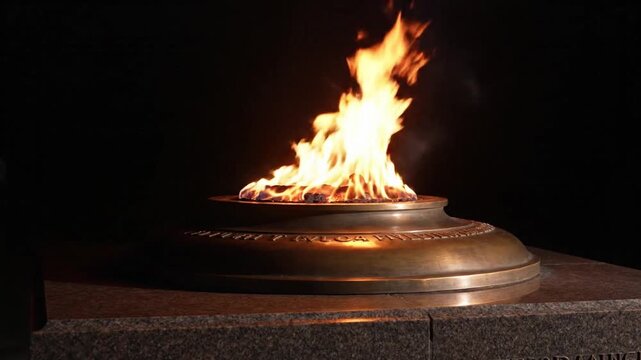 Bright orange and yellow flames flickering in a metal brazier on a stone pedestal against a dark background, representing eternal memory, remembrance, and paying tribute to fallen heroes