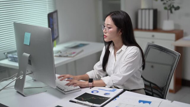Professional woman working on computer in modern office workspace with documents and charts