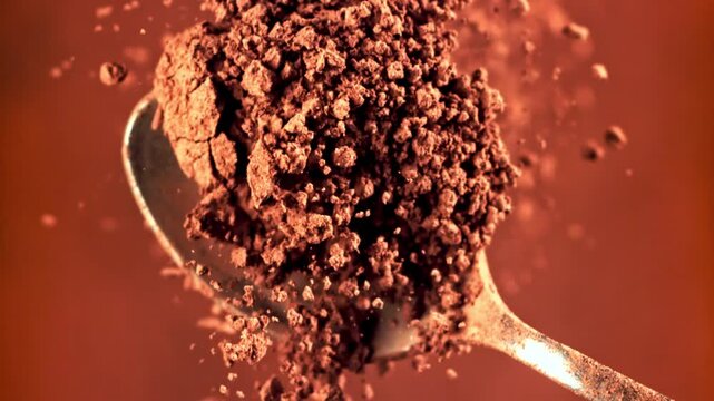 Close up shot of a metal spoon lifting ground aromatic brown cocoa chocolate powder with fine particles scattering against dark background