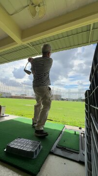 Horizontal video of a senior East Asian man with white hair wearing casual clothes and glasses practicing his swing at a golf driving range with an automatic tee system