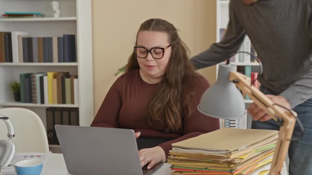 Woman typing on laptop while man leans over and rests hand on a stack of folders at desk in a building office with lamp and bookshelf visible; supportive collaboration.