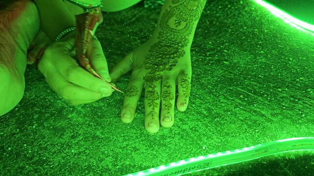 Close-up footage of a mehndi henna artist creating an intricate floral design on a woman's hand under decorative green LED lighting. Mehndi is an essential tradition during Indian weddings, festivals
