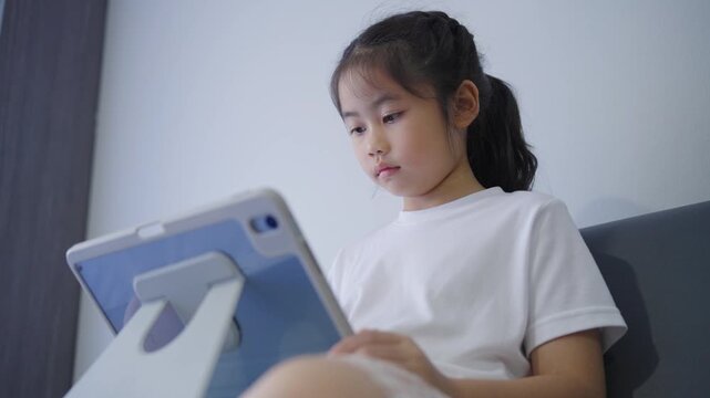 Young Asian girl sitting on a sofa using a tablet device with focused expression, casual white t-shirt, indoor home environment, modern interior design