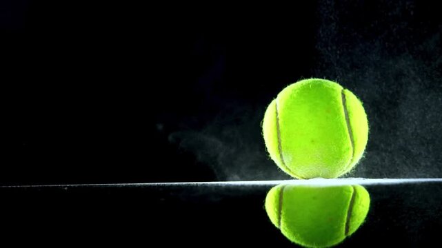 Macro shot of a bright yellow green tennis ball resting on a reflective glass surface with soft smoke rising on black background