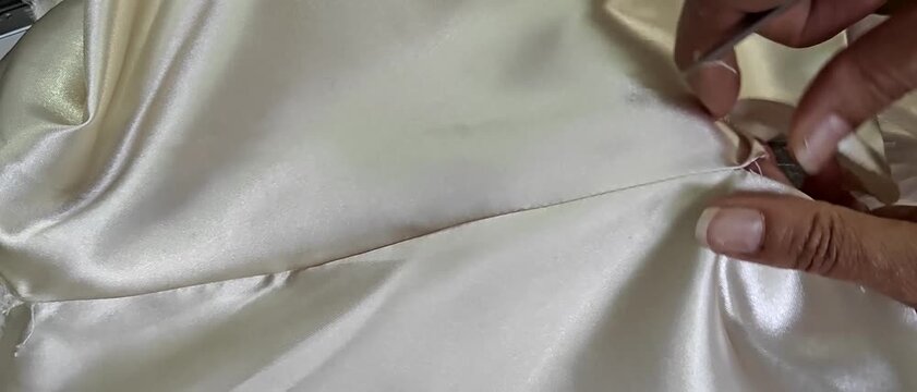 Hands removing stitches with small scissors from white satin fabric close up. Garment alteration, tailoring process, textile repair, delicate fabric work, sewing craftsmanship.