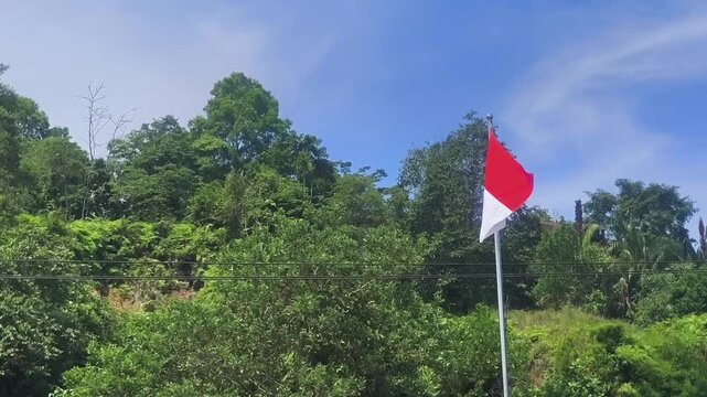 The Indonesian red and white flag waves beautifully on a flagpole with a lush green forest background.