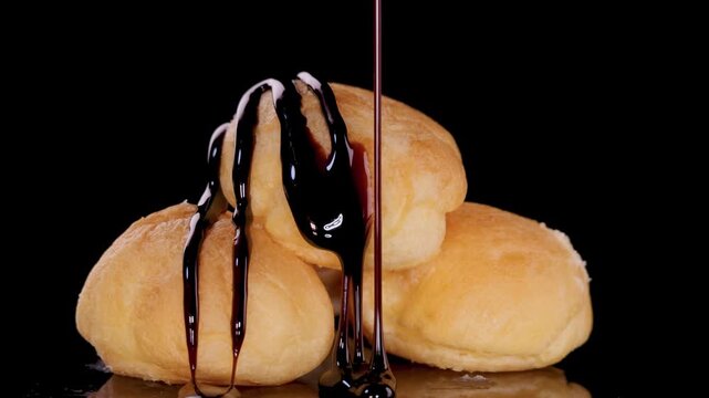 Close up shot of rich dark chocolate syrup being drizzled over delicious sweet cream puffs profiteroles against a black background indoors