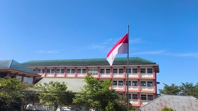 The large Indonesian red and white flag flutters under a bright blue sky in front of a school building.