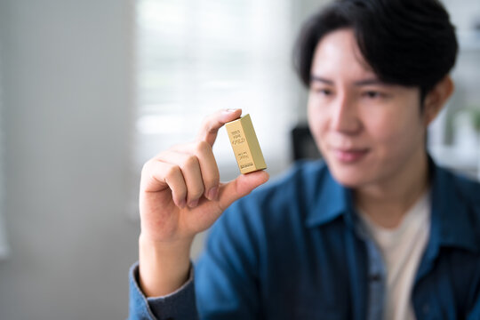 Young Asian investor examining gold bullion bar with smile in bright modern home office interior