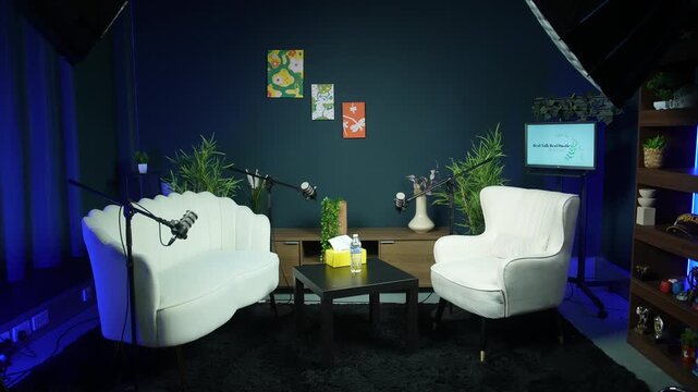 Empty professional podcast studio setup with microphones, white armchairs and modern interior decoration. Dark blue background wall, media broadcast recording and interview stage concept.