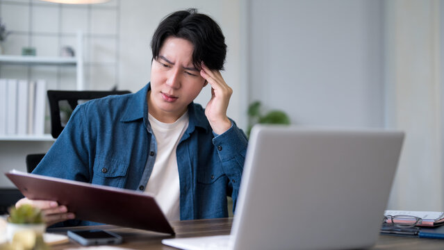 Young Asian man touching forehead experiencing work stress and headache while working with laptop and tablet at home office