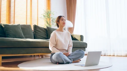 Fototapeten Lotusblume Happy young Asian woman practicing yoga and meditation at home sitting on floor in living room in lotus position with laptop  © Monster Ztudio