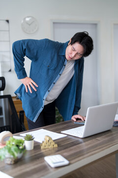 Young Asian man experiencing severe lower back pain while working on laptop at home office desk
