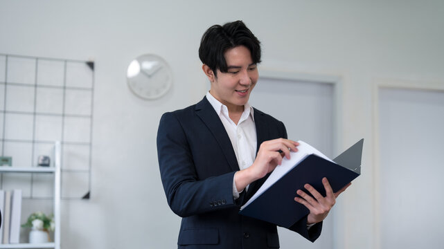 Young Asian businessman reviewing business documents in modern bright office workplace with confident smile