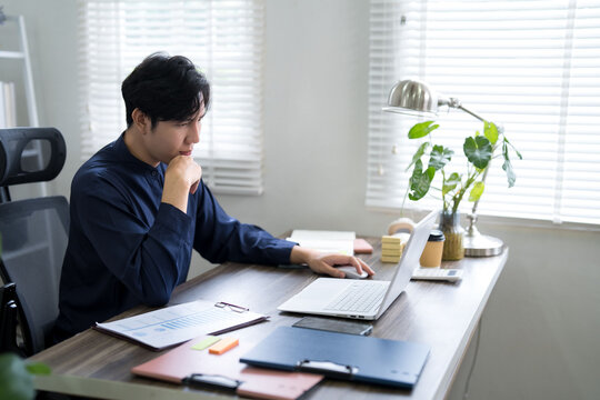 Young Asian businessman deep in thought working on laptop with financial charts at bright modern home office