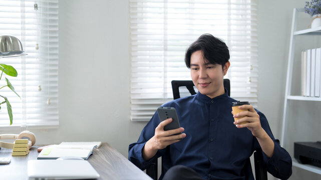 Young Asian professional checking smartphone while enjoying coffee break at bright modern home office workspace