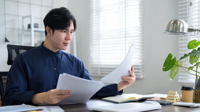 Young Asian business professional reviewing contract documents in bright modern home office workspace