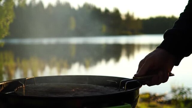 Man grilling food by lake at sunset, steam rising from barbecue