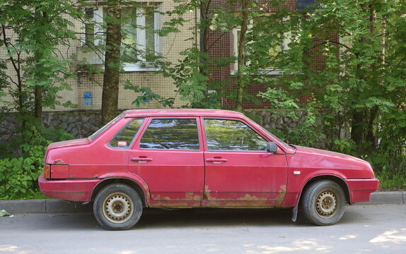 A red, old, rusty car is parked near a lawn in front of a residential building, Krzhizhanovsky Street, Saint Petersburg, Russia, June 21, 2026