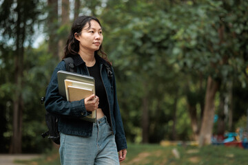 Fototapeta premium Young Asian Female Student Walking Outdoors with Laptop and Notebooks Wearing Denim Jacket and Backpack