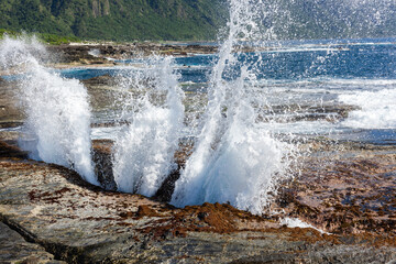 Naklejka premium Shitiping coastal blowholes in Hualien, Taiwan. Ocean water splashes through rock formations. Rocky shoreline with green mountains in the distance. Pacific Ocean waves crashing.