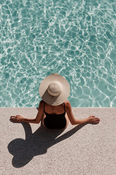 Looking down from above, a woman wearing a large straw hat relaxes on the edge of a rippling turquoise swimming pool.