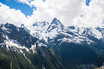 Fototapeta premium Snow-covered mountain peaks rise against a blue sky with clouds, surrounded by green valleys and rocky slopes, creating a stunning natural landscape