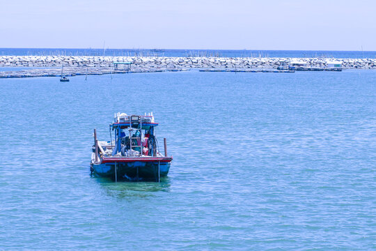 Small traditional fishing boat floating in the calm sea at Ang Sila, Chonburi, Thailand, with coastal aquaculture, breakwater, and blue ocean under a clear daytime sky.