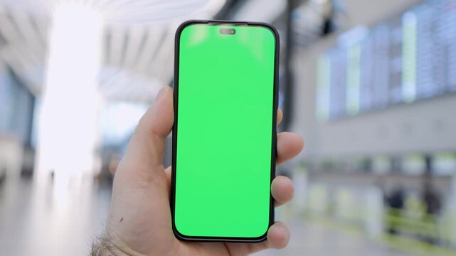 Hand holds phone with green screen at airport. Background shows blurred terminal and flight boards. Focus remains on phone, ready for custom content. Ideal for travel, technology, or app demos