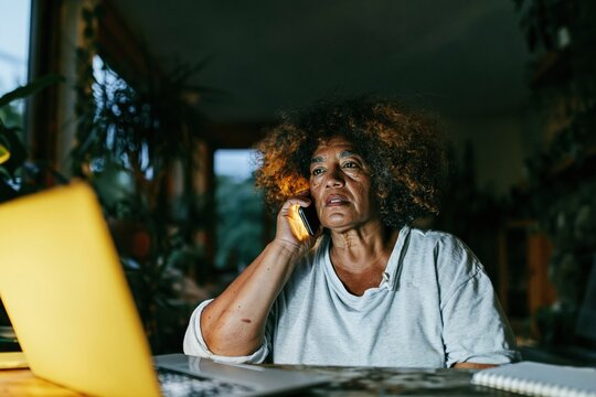 Focused woman talking on her smartphone while working at a laptop in a moody home office, for remote work and freelancing themes
