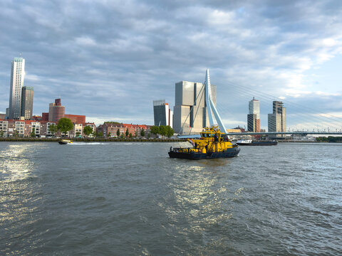 Rotterdam, Netherlands skyline with Erasmus Bridge, modern buildings and a yellow tugboat on river, cloudy sky.