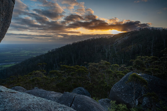 View of Castle Rock at sunset with granite boulders, dense forest slopes, and golden sun glowing behind clouds over the horizon in Porongurup, Western Australia, Australia.