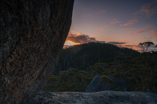 View of Castle Rock at sunset with large granite boulders in the foreground and forest-covered hills under a colorful sky in Porongurup, Western Australia, Australia.