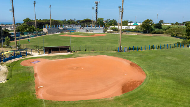 Wide aerial view of a professional baseball stadium under a clear blue sky, showing the green grass outfield, detailed red dirt infield, dugouts, tall stadium light poles, and empty spectator stands.