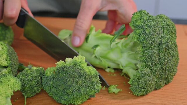 Hands cutting fresh broccoli with knife on wooden board during food preparation. Healthy nutrition concept featuring green vegetable ingredients for diet planning, wellness goals and home cooking.