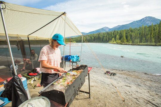 Smiling woman prepares food outdoors while camping by a mountain river