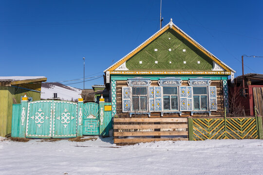 View of a traditional wooden house with ornate blue shutters and a green gable roof in a snowy landscape in Baikal'skoe, Buryatia, Russia.