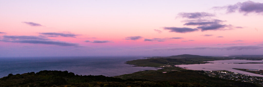A panoramic view of a pink and purple sunset over ocean with land in foreground and purple clouds