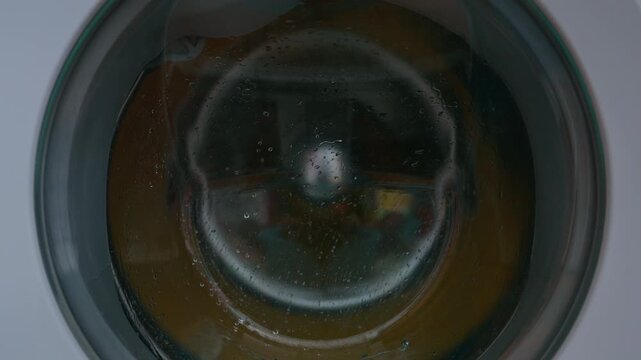 Clothing and soapy water spin around in machine. Washing machine door. Front loader