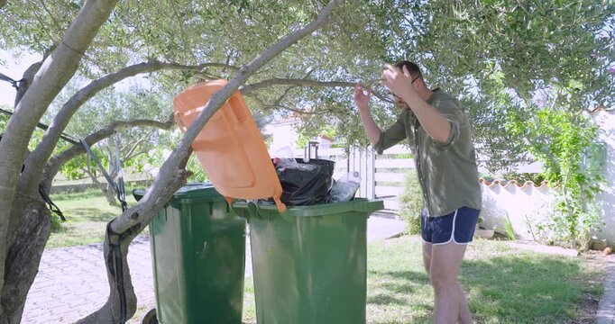 A frustrated man stands beside an overflowing garbage container in a backyard garden, expressing annoyance while dealing with household waste and an overfilled trash bin outdoors.
